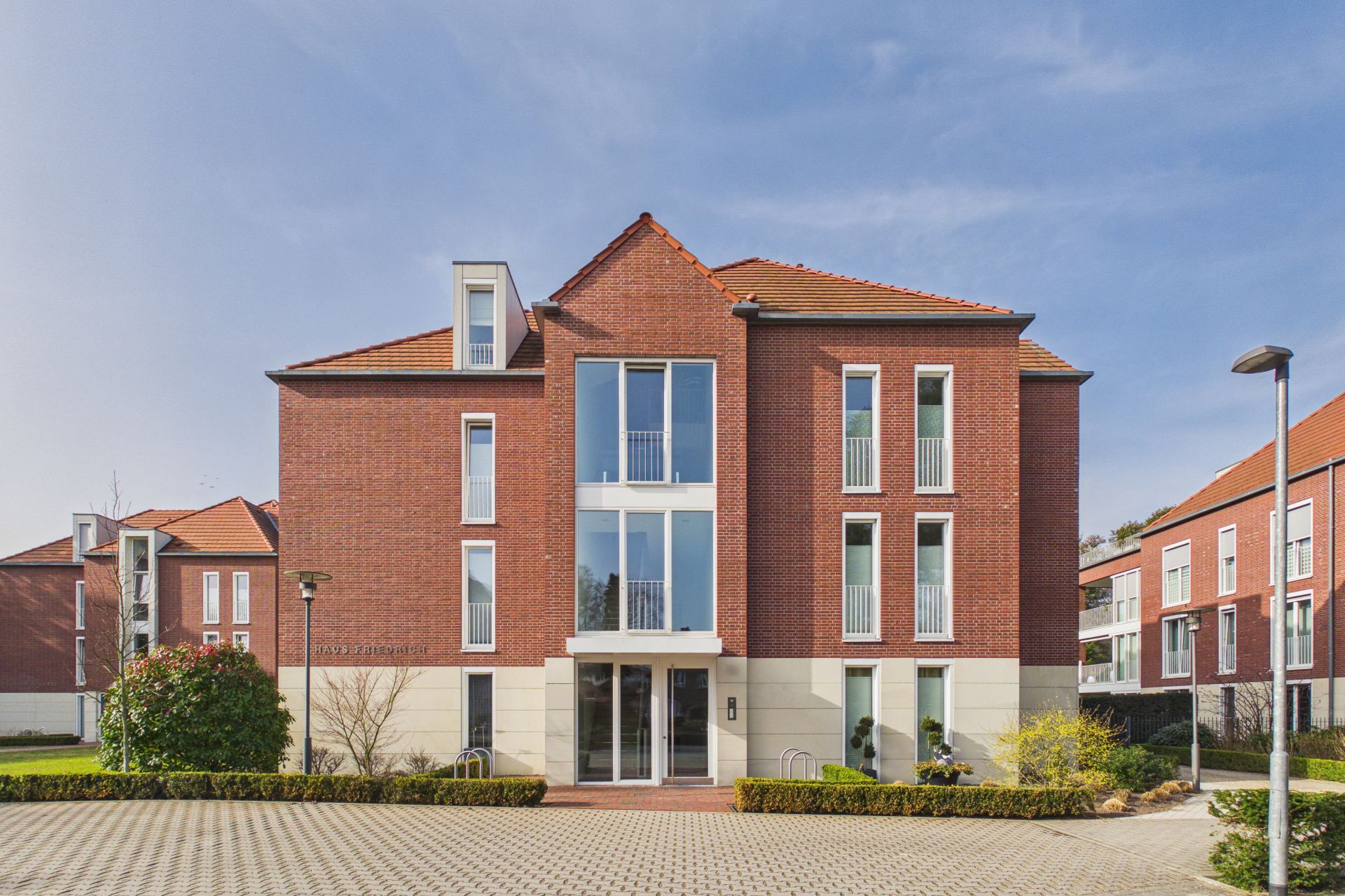 Modernes Mehrfamilienhaus mit roten Ziegeln und großen Fenstern, Parkplatz vorne.
