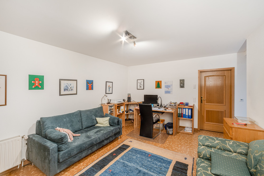 Wohnzimmer mit Sofa, Schreibtisch, Holzregalen, Teppich und mehreren Bildern an der Wand.