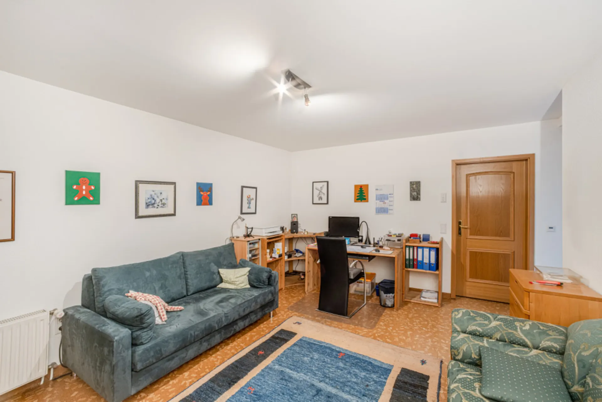 Wohnzimmer mit Sofa, Schreibtisch, Holzregalen, Teppich und mehreren Bildern an der Wand.