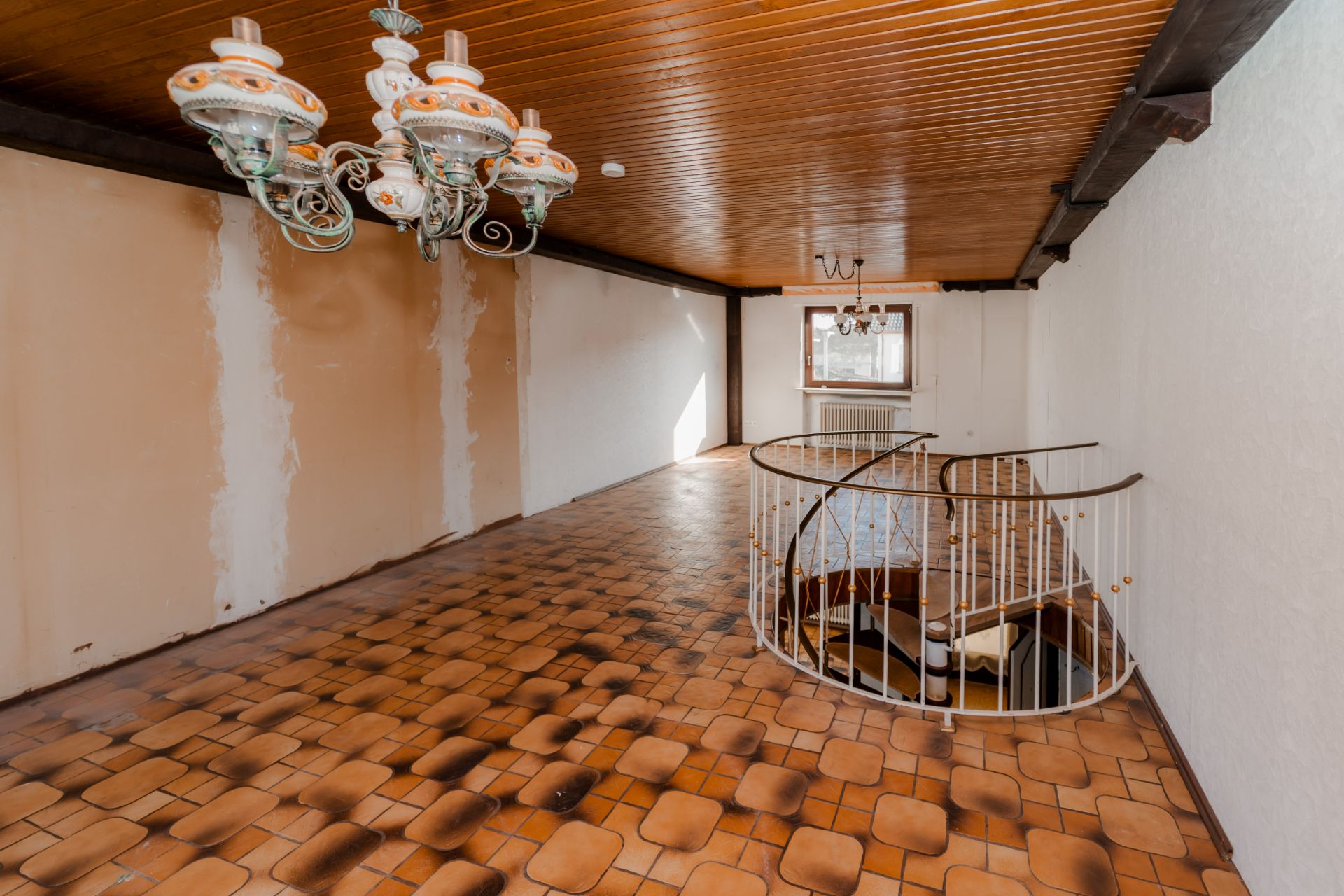 Leerer Raum mit braunen Fliesen, Holzdecke, Kronleuchter und Wendeltreppe nach unten.
