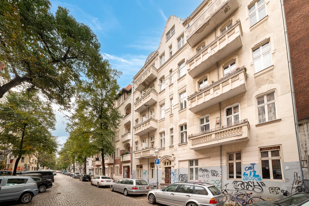 Mehrstöckiges Wohnhaus mit Balkonen an einer Baumgesäumten Straße mit parkenden Autos.