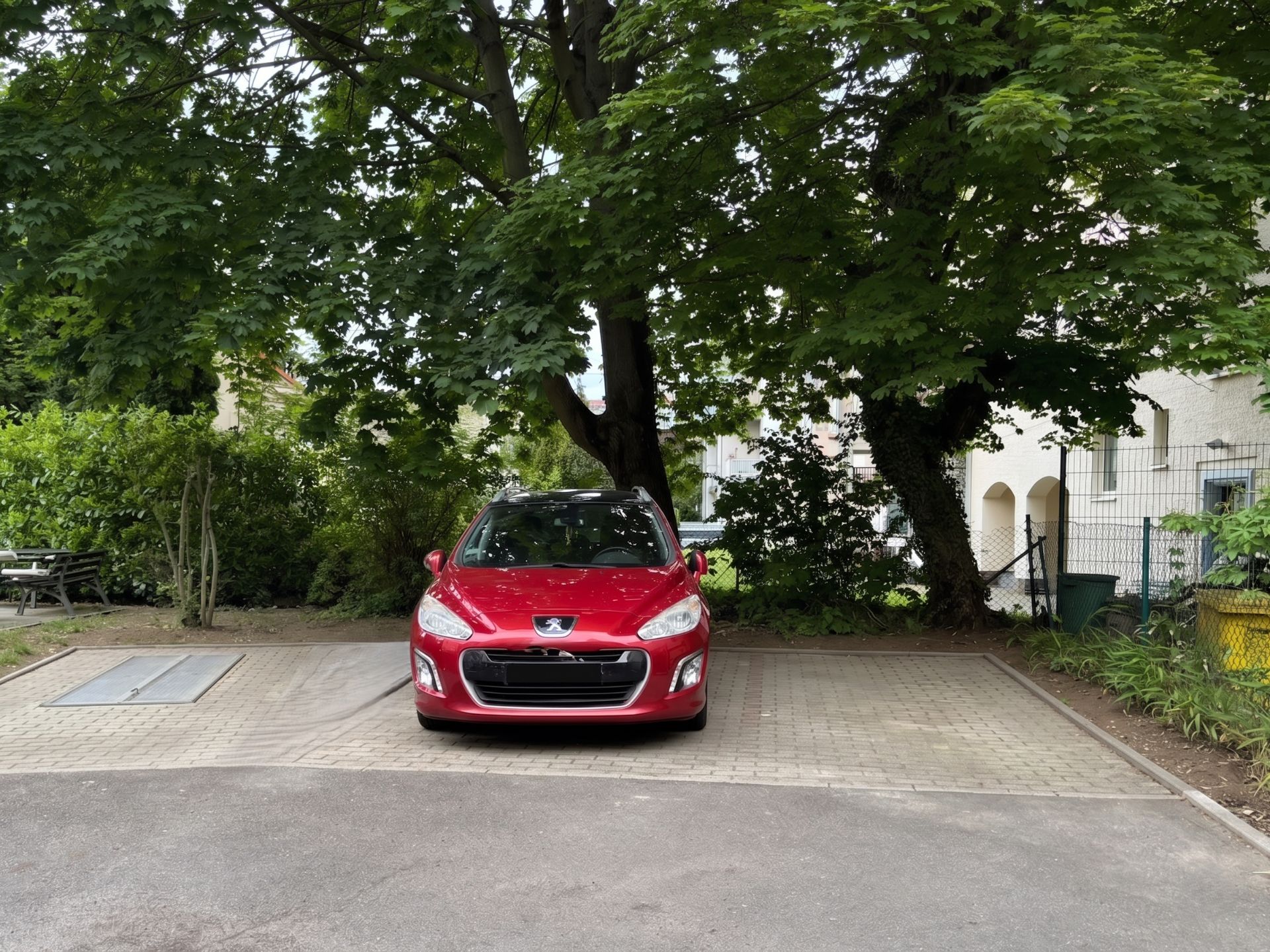 Roter Pkw steht auf einem Parkplatz unter einem großen Baum, im Hintergrund Garten und Wohngebäude.