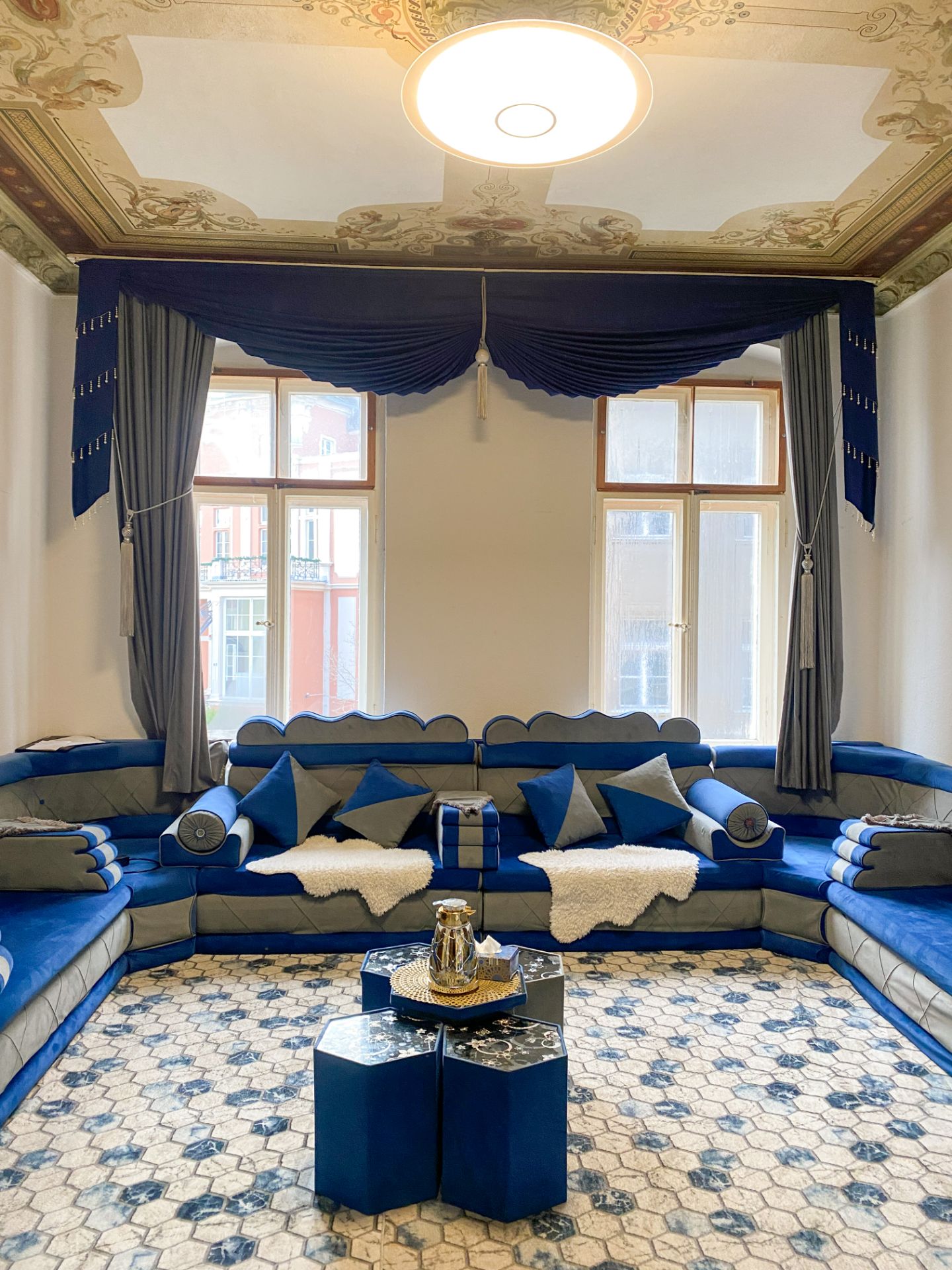 Wohnzimmer mit blauer Sofalandschaft, gemustertem Teppich und verzierten Vorhängen vor zwei Fenstern.