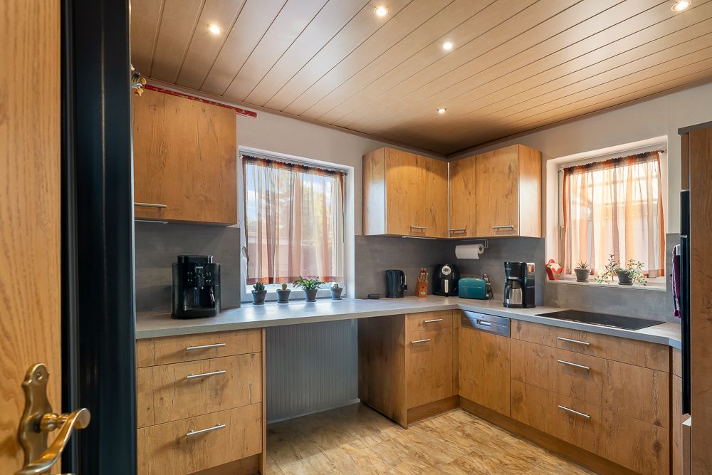 Moderne Küche mit Holzfronten, Geräten, zwei Fenstern und kleinen Pflanzen auf der Arbeitsfläche.