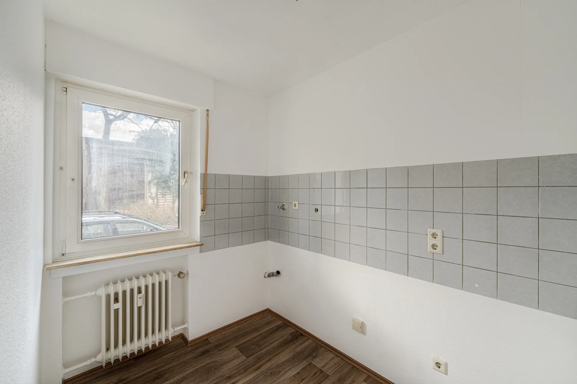 Leerer Raum mit Fenster, grauen Fliesen an der Wand und Heizkörper unter dem Fenster.