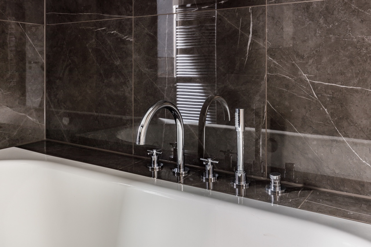 Weiße Badewanne mit modernen Chromarmaturen vor dunklen Marmorfliesen.