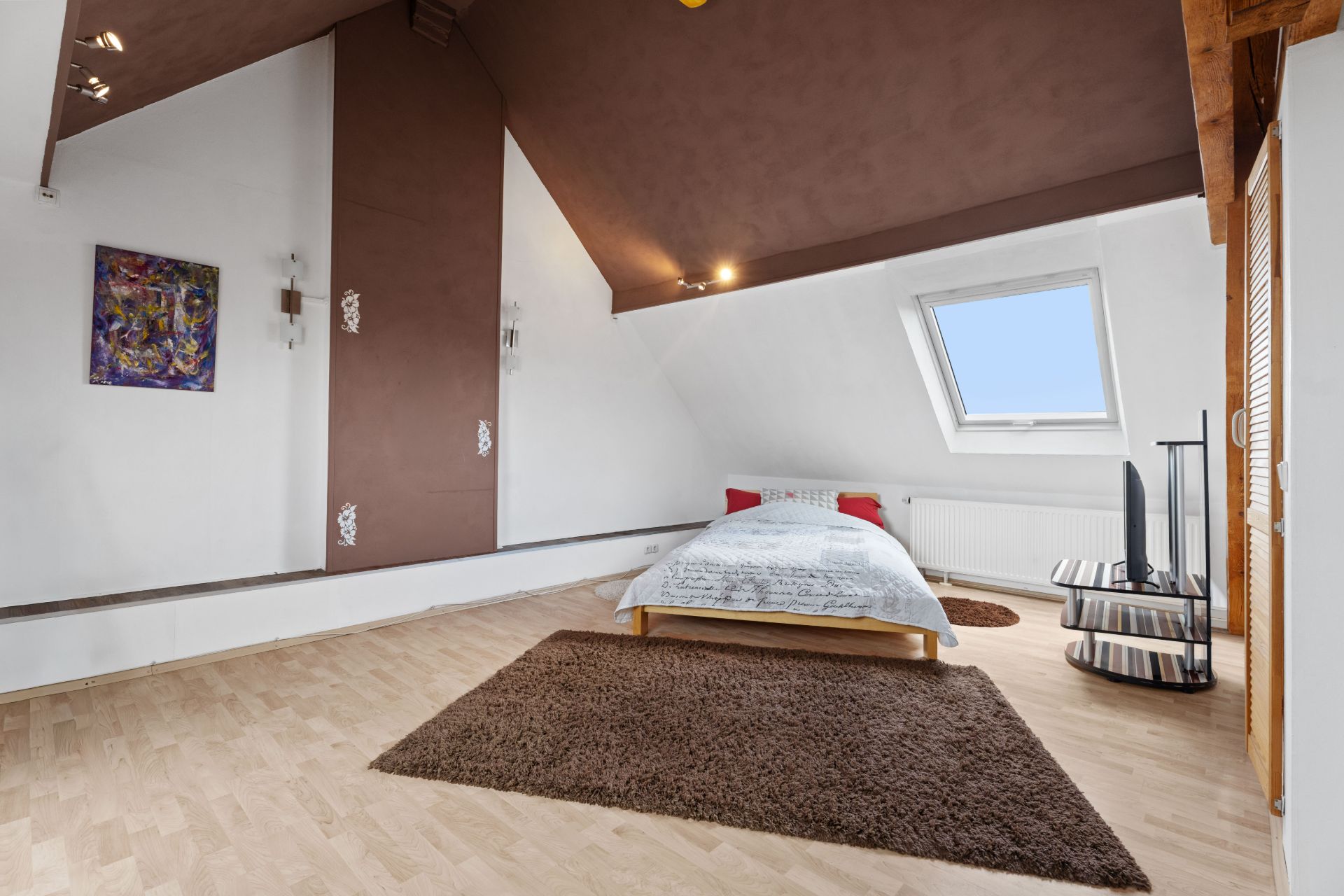 Schlafzimmer mit Dachschräge, Bett, Teppich, Fenster und TV auf hellem Holzboden.