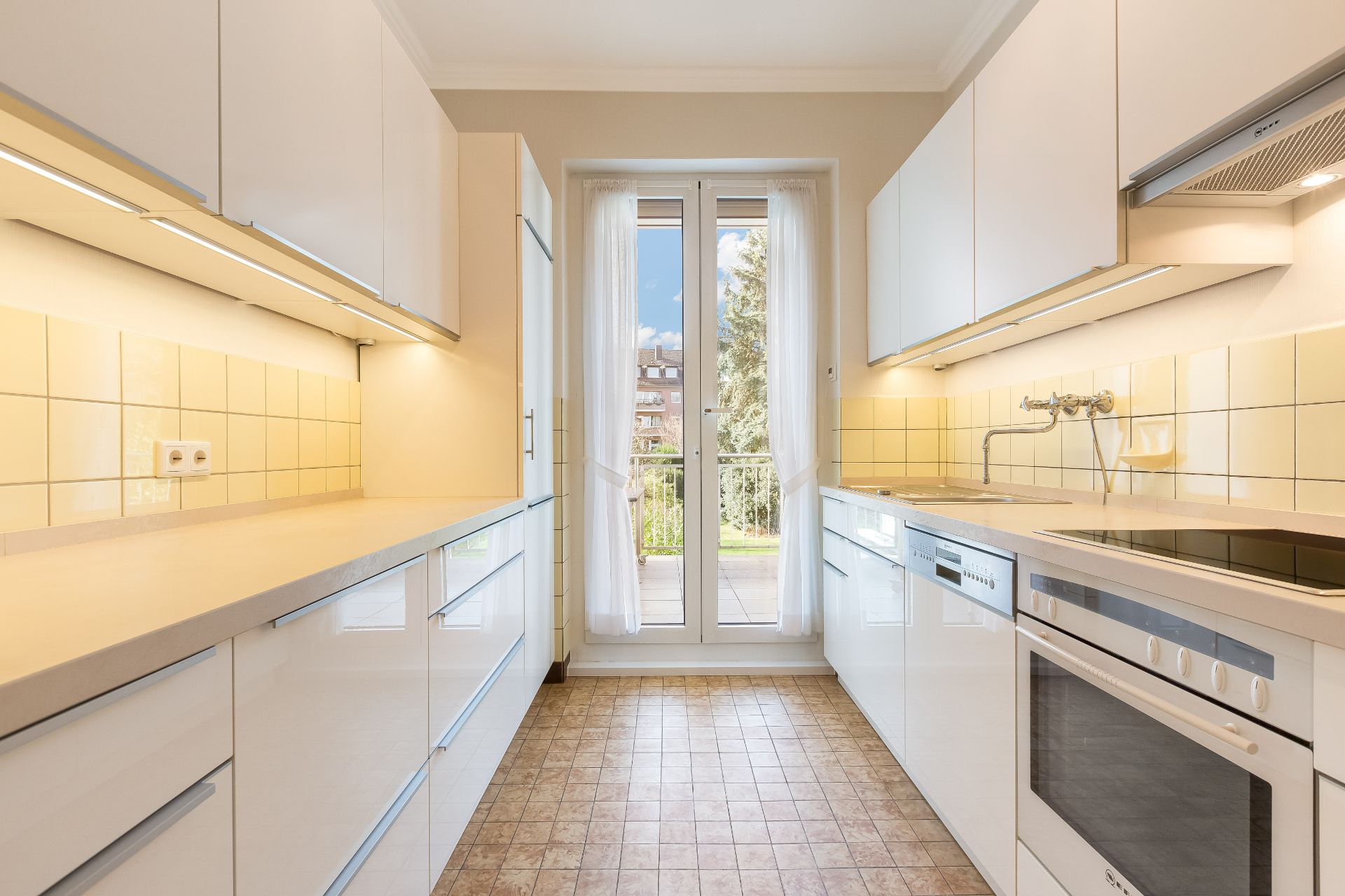 Moderne, helle Einbauküche mit weißen Schränken, Fliesenboden und Zugang zum Balkon.