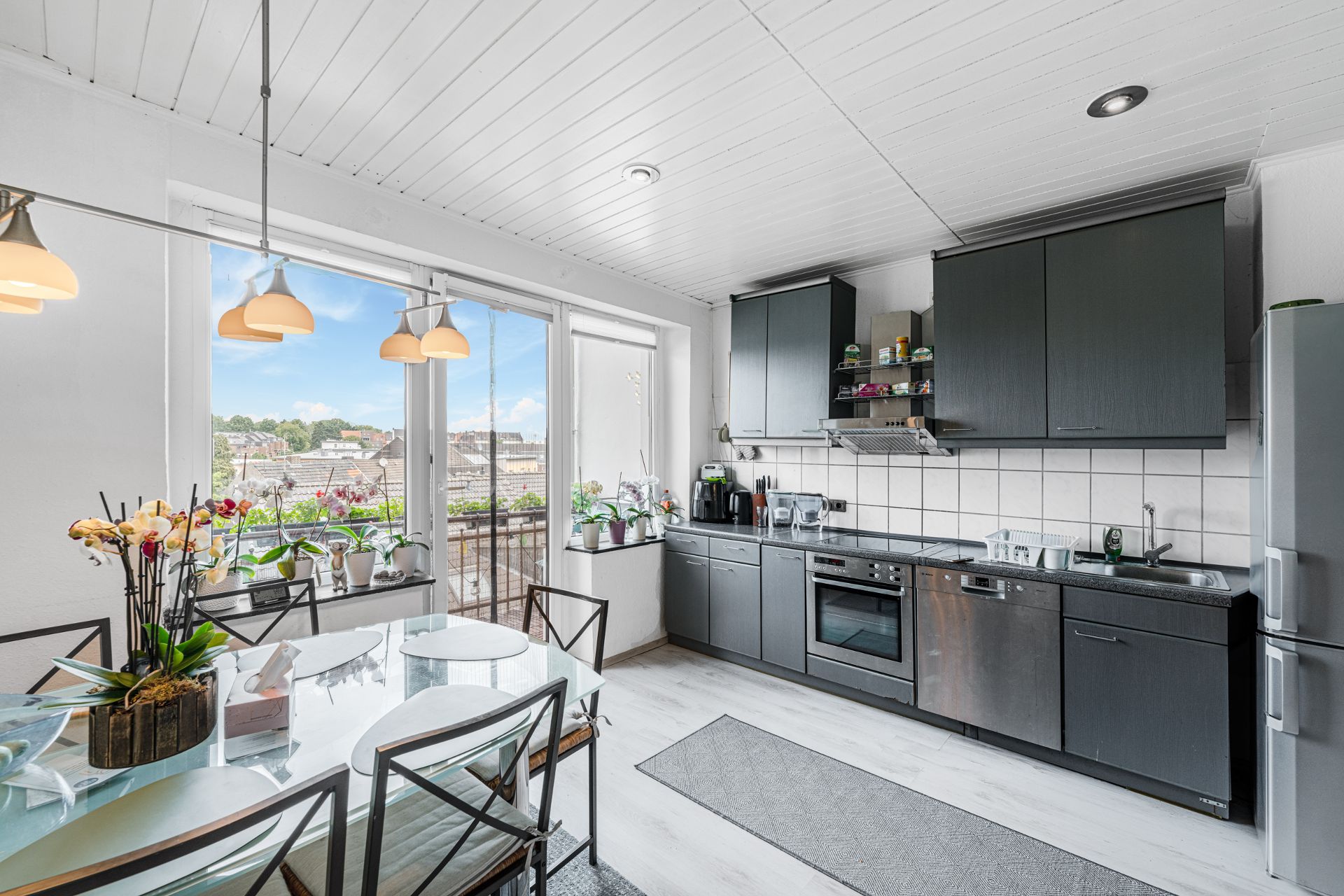 Moderne Küche mit grauen Schränken, Esstisch aus Glas und großen Fenstern mit Blick auf einen Balkon.
