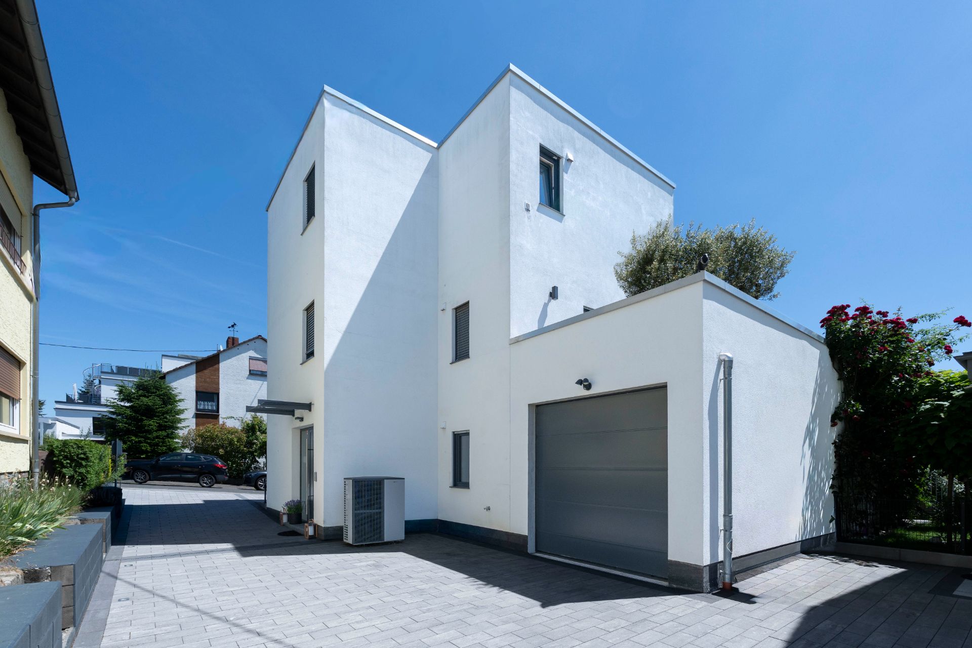 Modernes, weißes Wohnhaus mit Flachdach und Garage, umgeben von Einfahrt und etwas Grün.