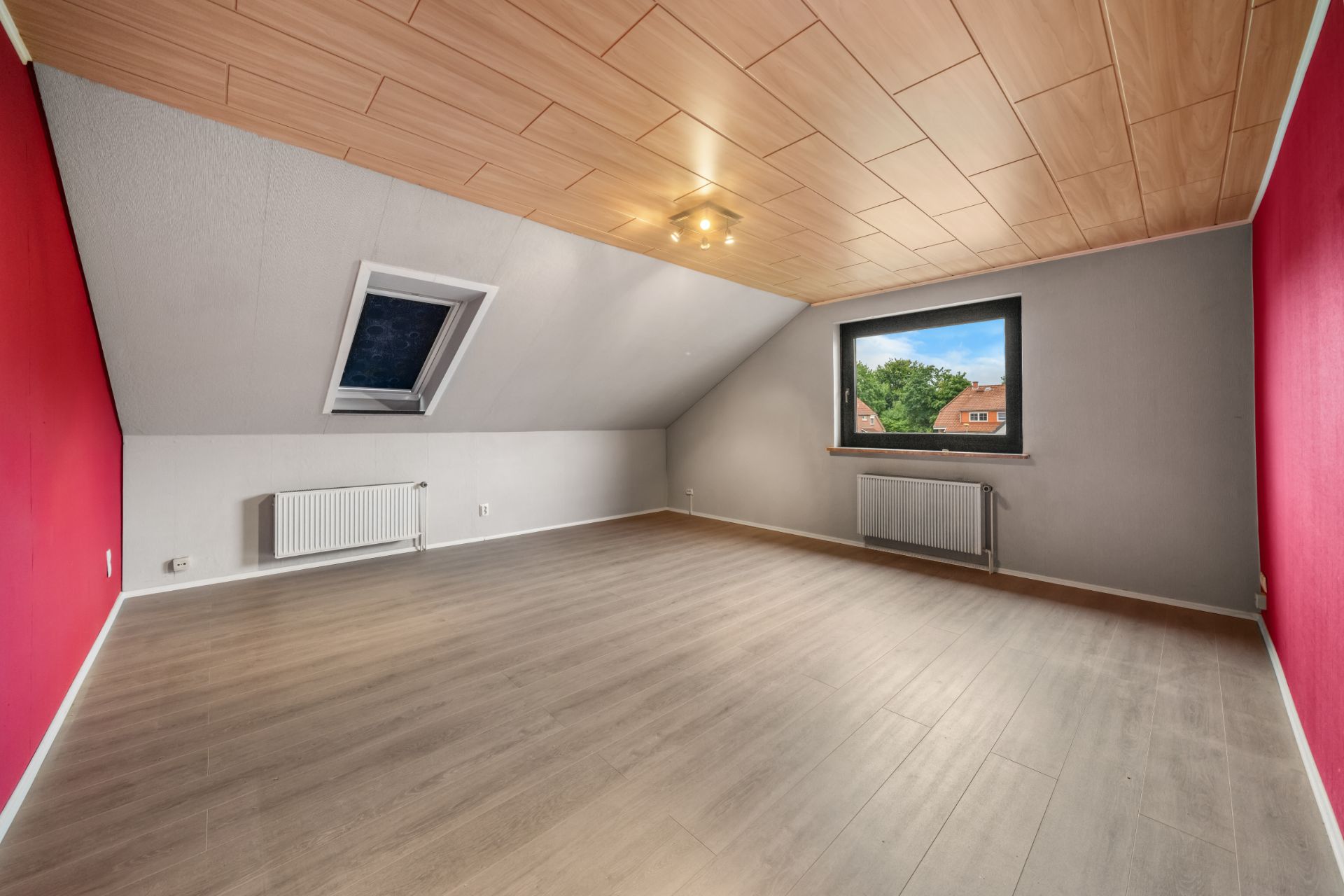 Leeres Dachgeschosszimmer mit Holzdecke, Dachfenster und normalem Fenster, zwei Heizkörpern und Holzfußboden.
