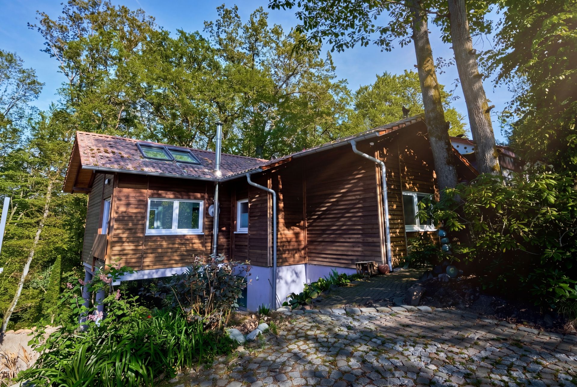 Holzhaus mit großen Fenstern und Dachfenstern im Grünen, davor gepflasterter Weg.