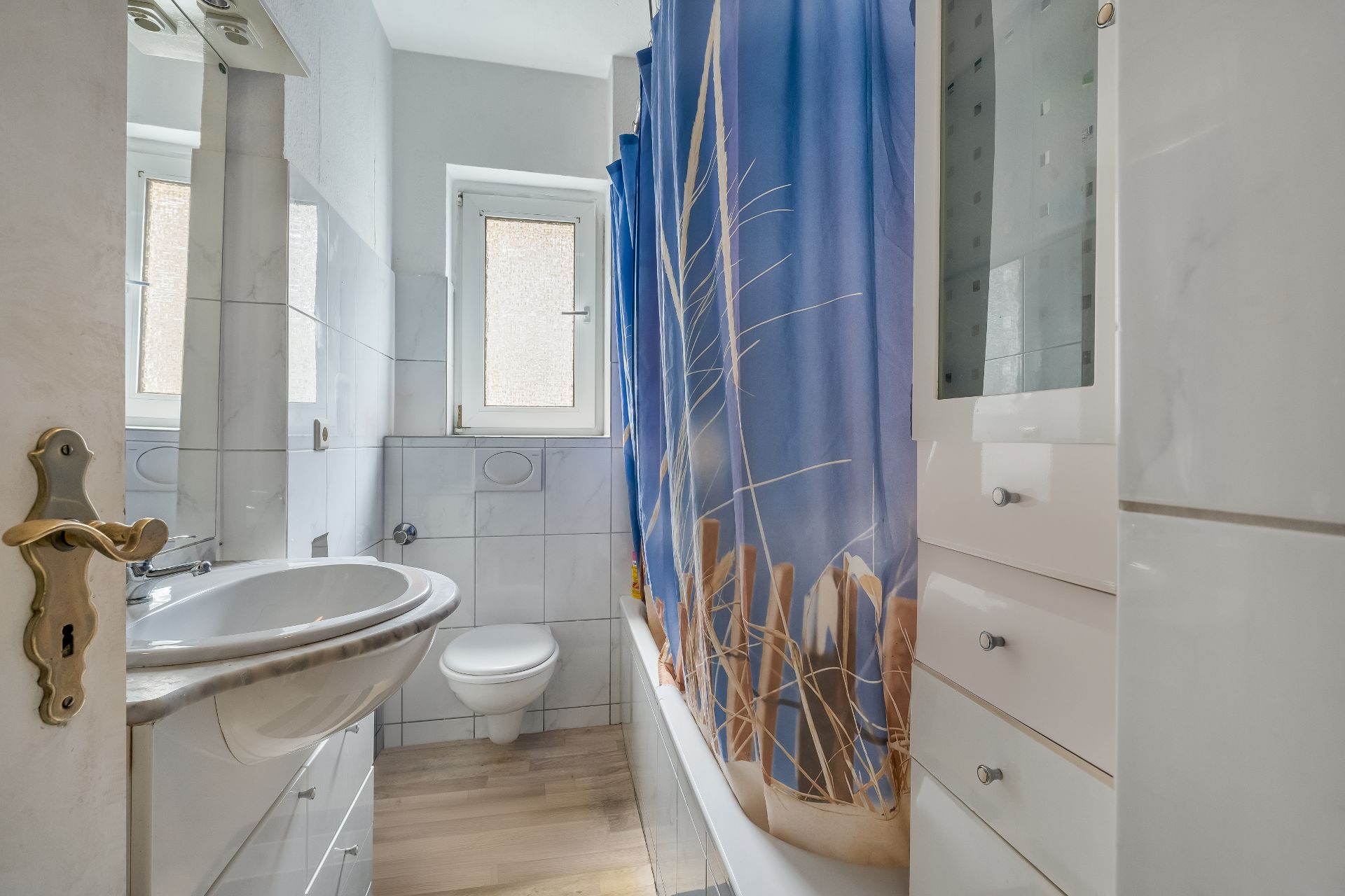Kleines, helles Badezimmer mit Waschbecken, WC, Dusche mit Duschvorhang und Fenster.
