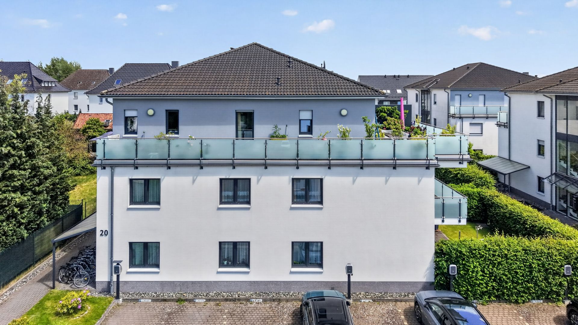 Modernes Wohnhaus mit Balkon, Flachdach und mehreren Fenstern, davor Parkplätze und Fahrräder.