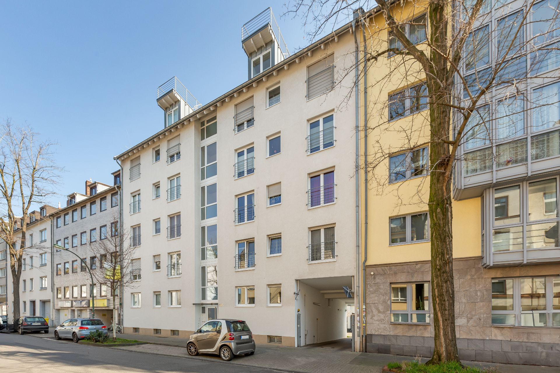 Fünfgeschossiges Wohnhaus mit weißer Fassade, mehreren Fenstern und geparkten Autos davor.