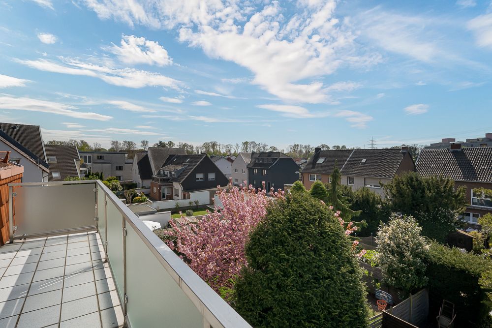 Blick von einem Balkon auf Nachbarhäuser, Bäume und blühende Sträucher unter blauem Himmel.