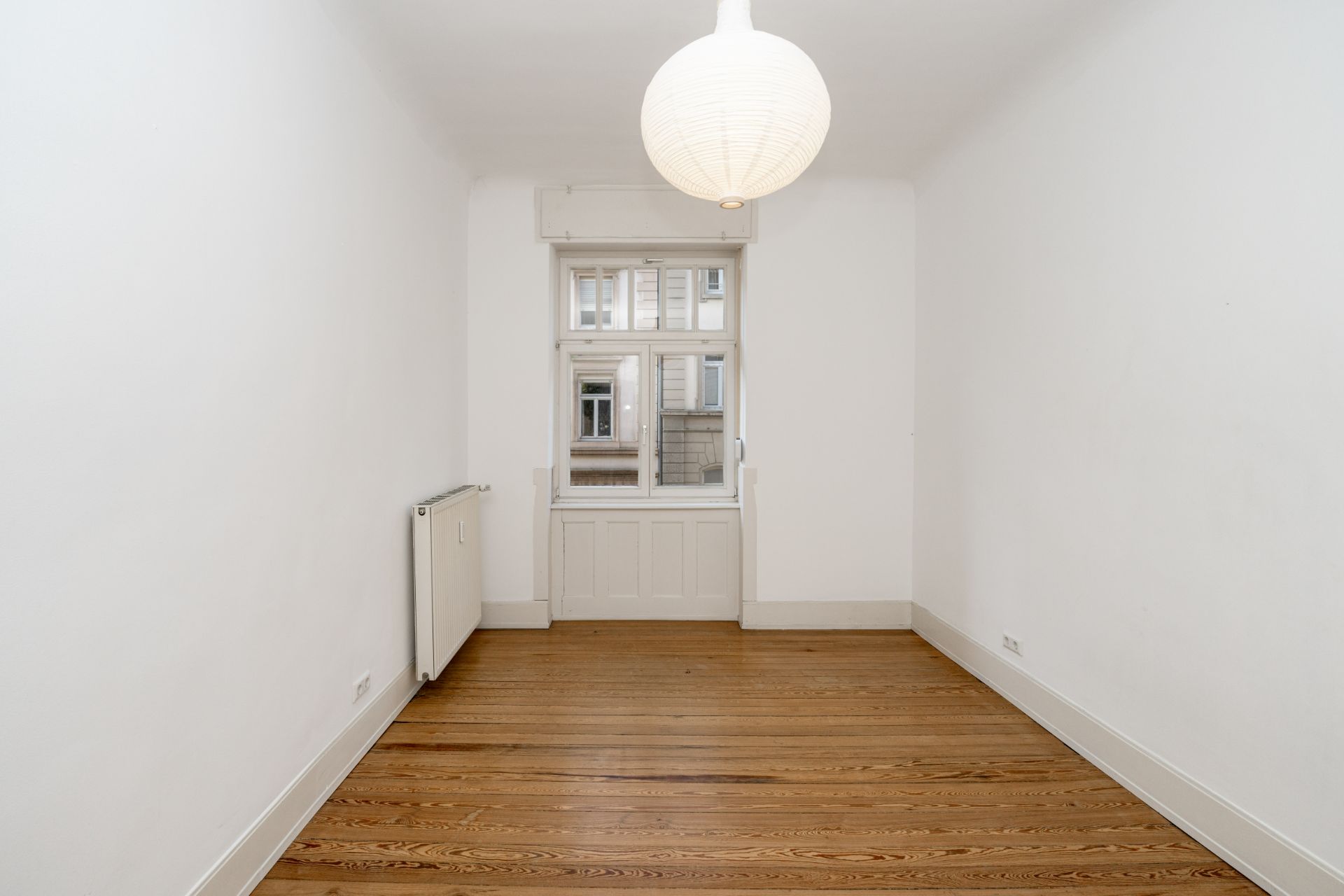 Leerer Raum mit Holzboden, weißer Wand, Fenster und runder Deckenleuchte.