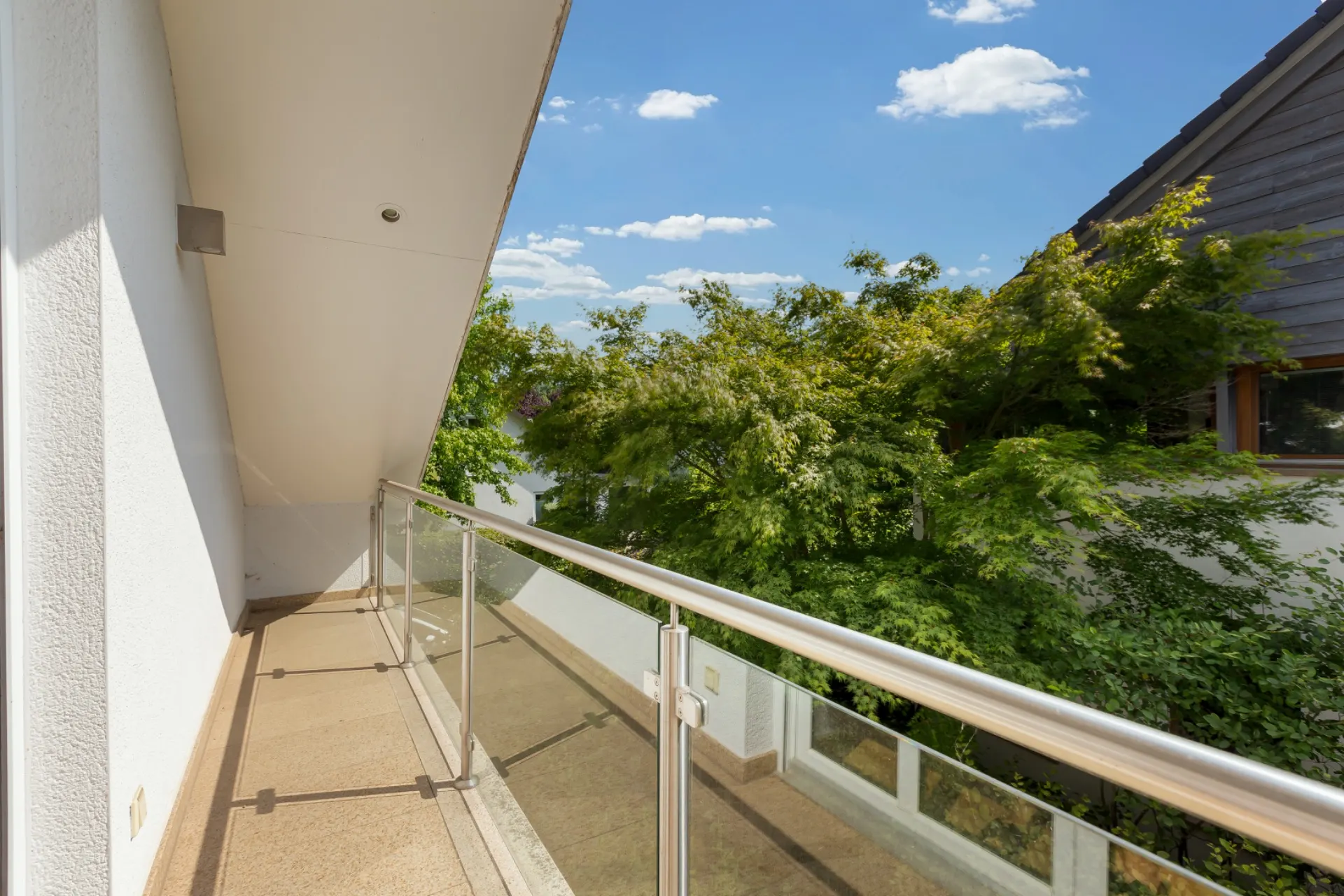 Balkon mit Glasgeländer und Blick auf grüne Bäume bei sonnigem Wetter.