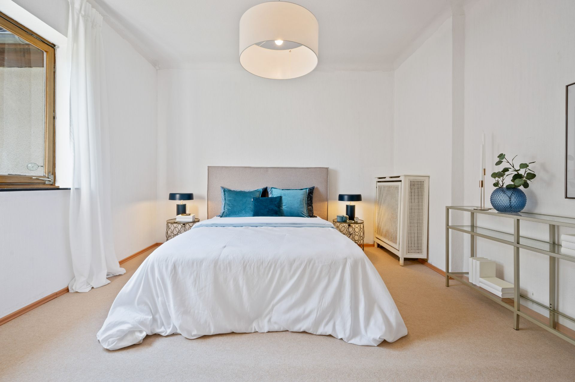 Helles Schlafzimmer mit Doppelbett, blauen Kissen, zwei Nachttischen und großer Deckenleuchte.