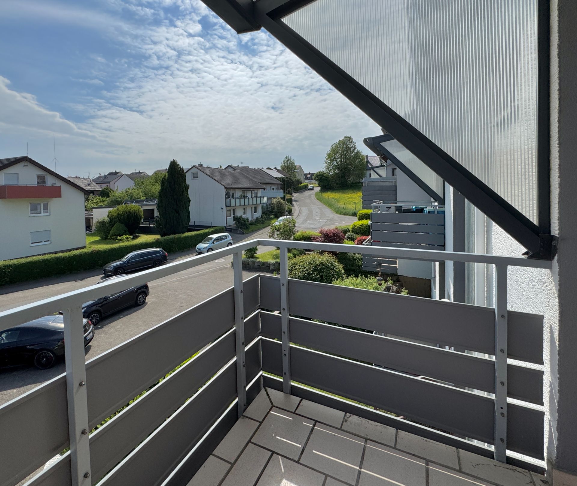 Balkon mit grauem Geländer und Blick auf eine ruhige Wohnstraße und Nachbarhäuser.