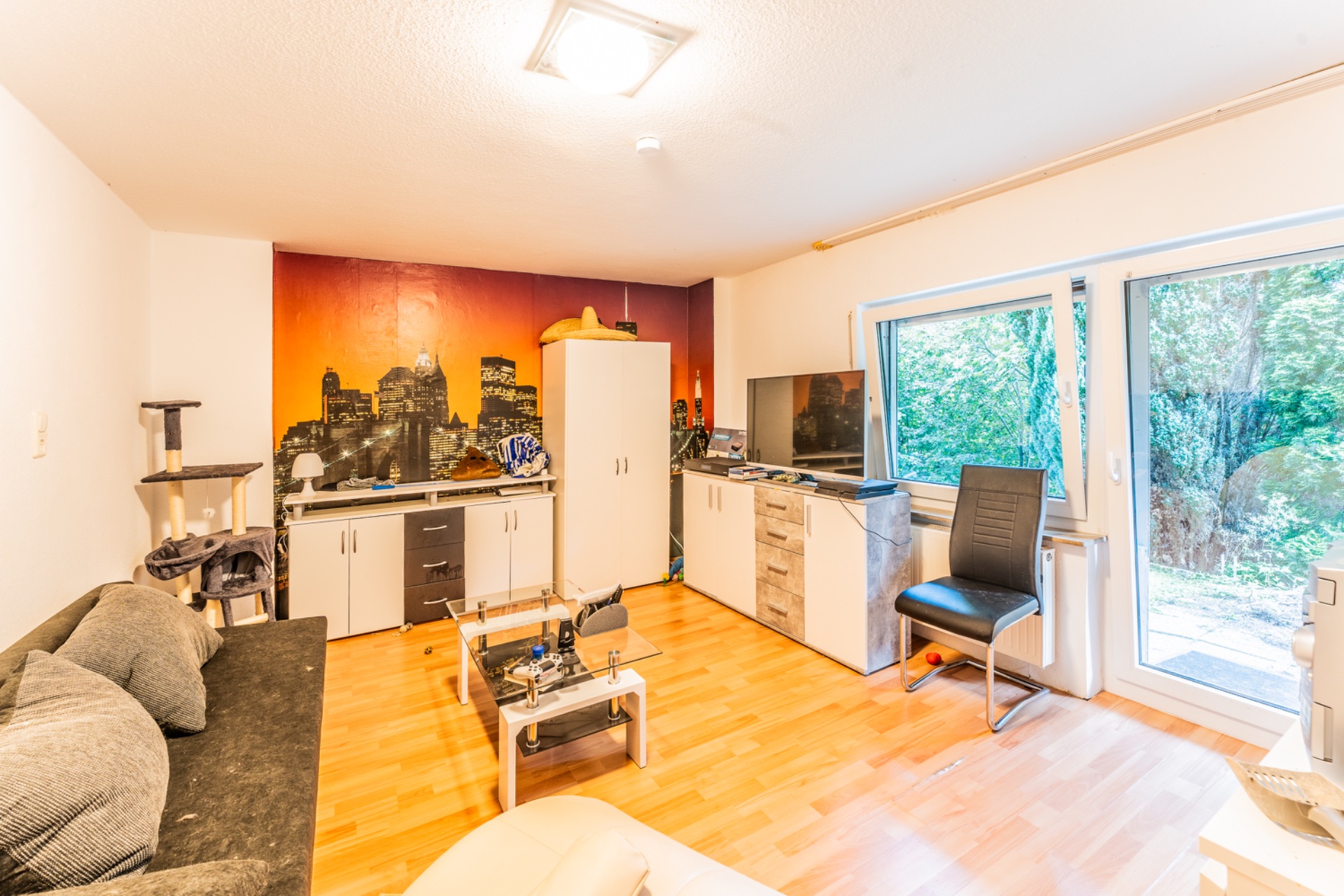 Helles Wohnzimmer mit Sofa, Couchtisch, Kommode, Fernseher, Stuhl und Blick ins Grüne.