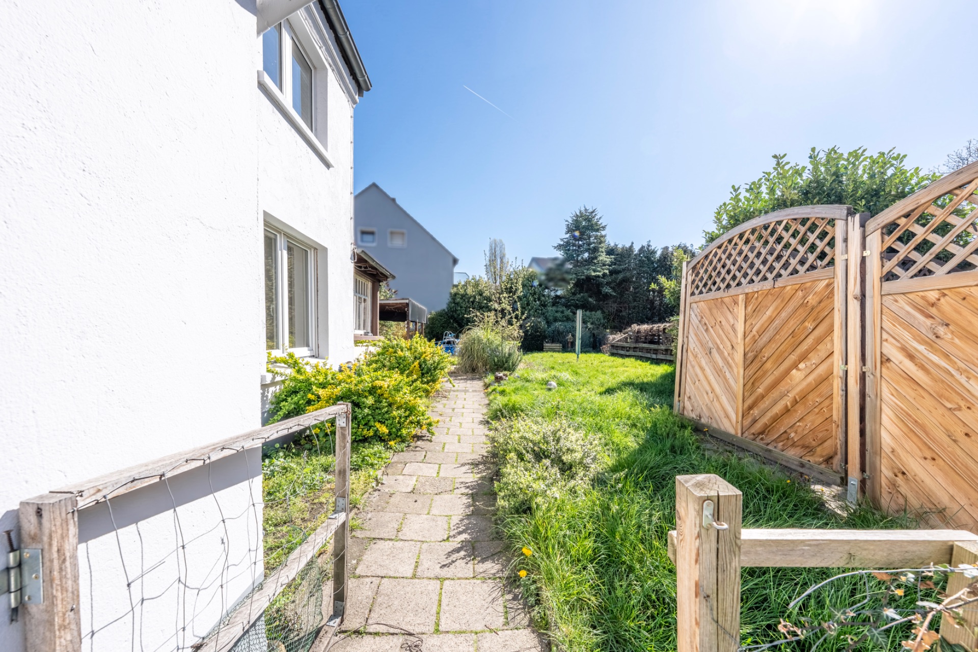 Gepflasterter Weg neben einem weißen Haus, rechts Holzzaun und grüne Wiese bei Sonnenschein.