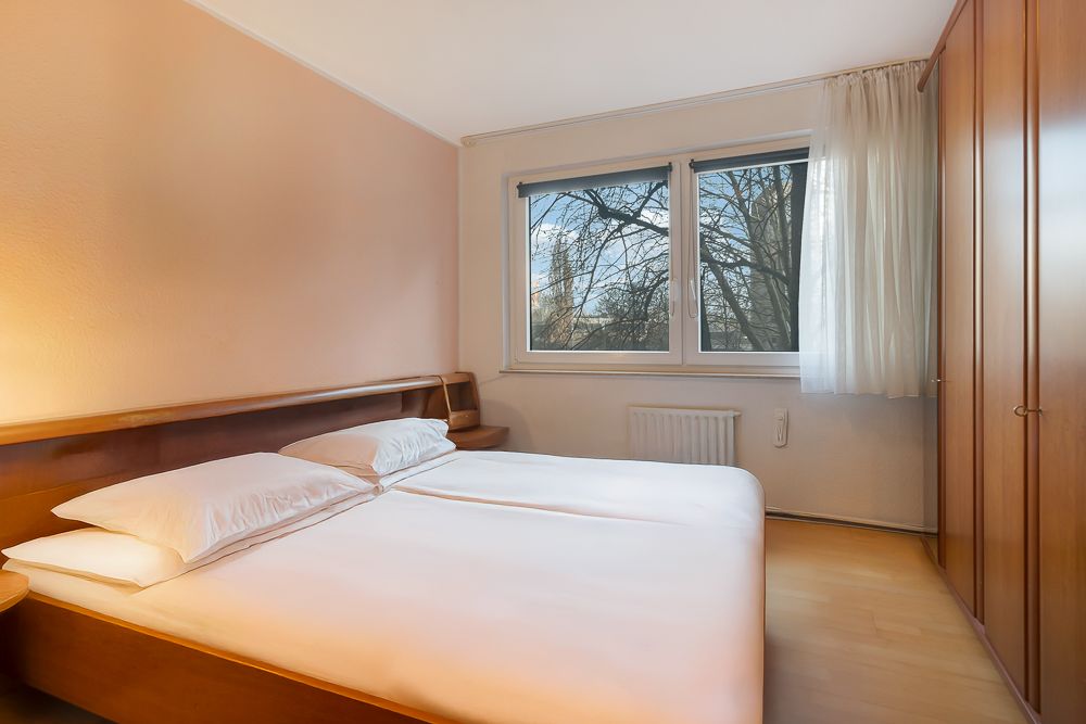 Schlafzimmer mit Doppelbett, zwei Fenstern und einem großen Kleiderschrank aus Holz.