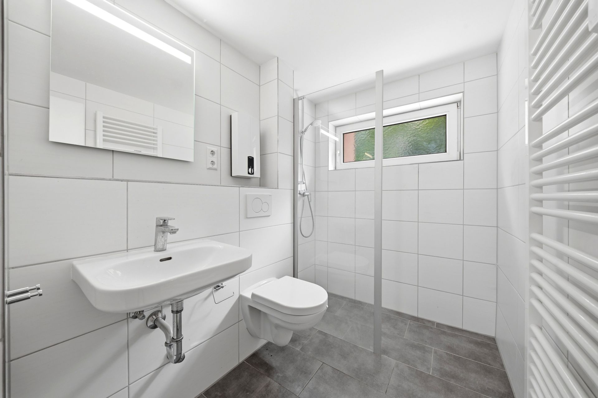 Modernes, weiß gefliestes Badezimmer mit Waschbecken, WC, bodengleicher Dusche und Fenster.