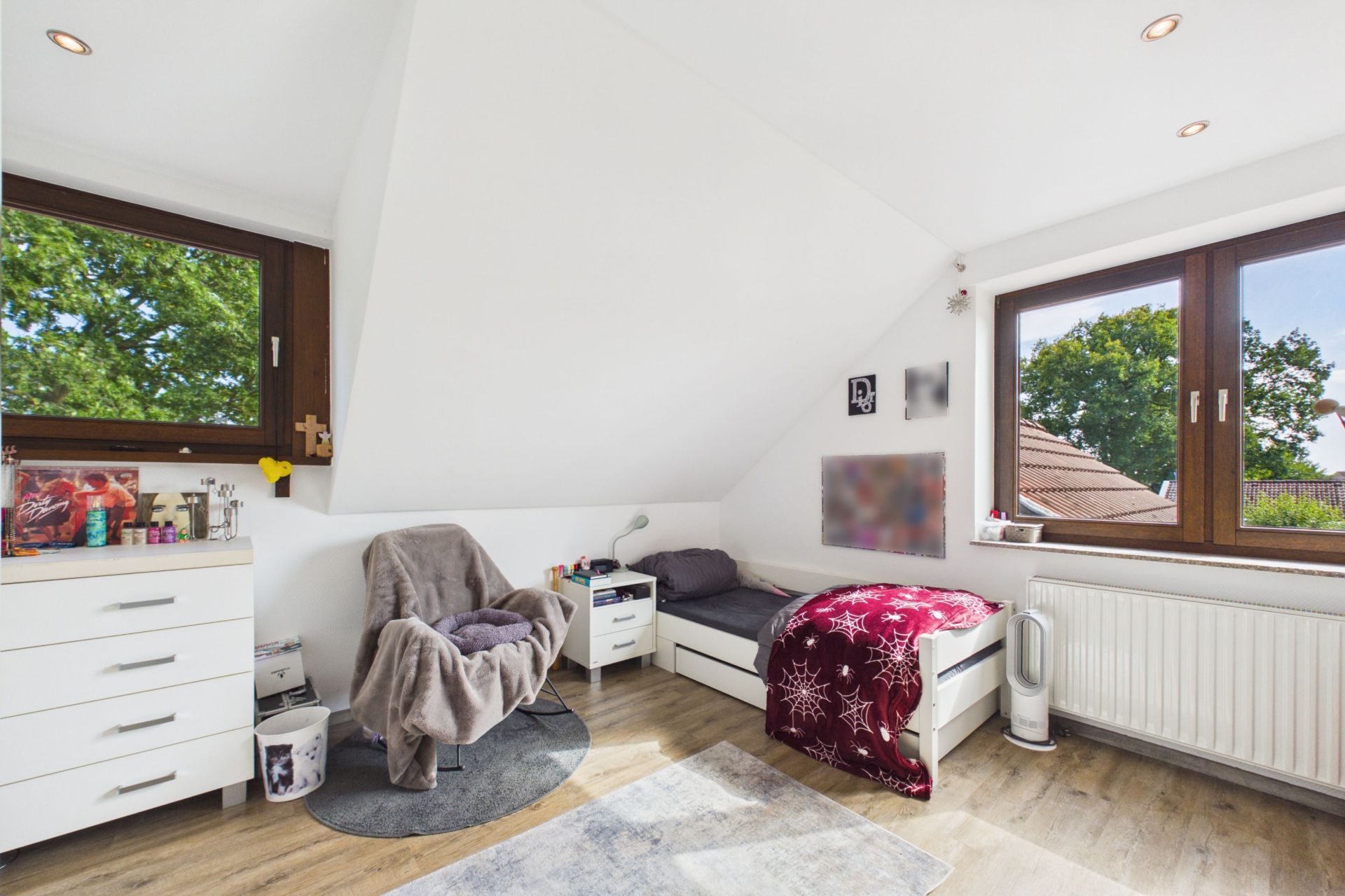 Helles Schlafzimmer mit Einzelbett, Sessel, Kommode und zwei großen Fenstern.