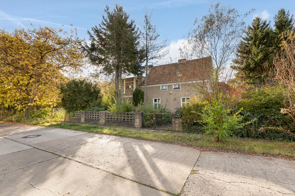 Zweistöckiges Wohnhaus mit rotem Dach, umgeben von Bäumen und Sträuchern, Blick von der Straße.
