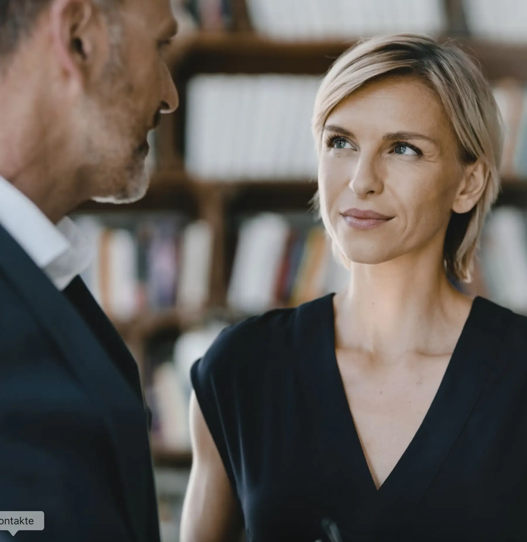Zwei Personen unterhalten sich in einem Raum mit Bücherregal im Hintergrund.