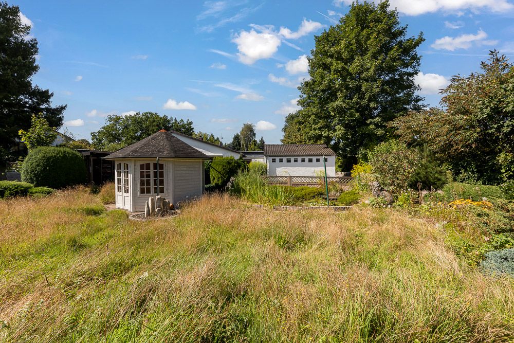 Garten mit hohem Gras, einem kleinen Pavillon und Bäumen im Hintergrund.