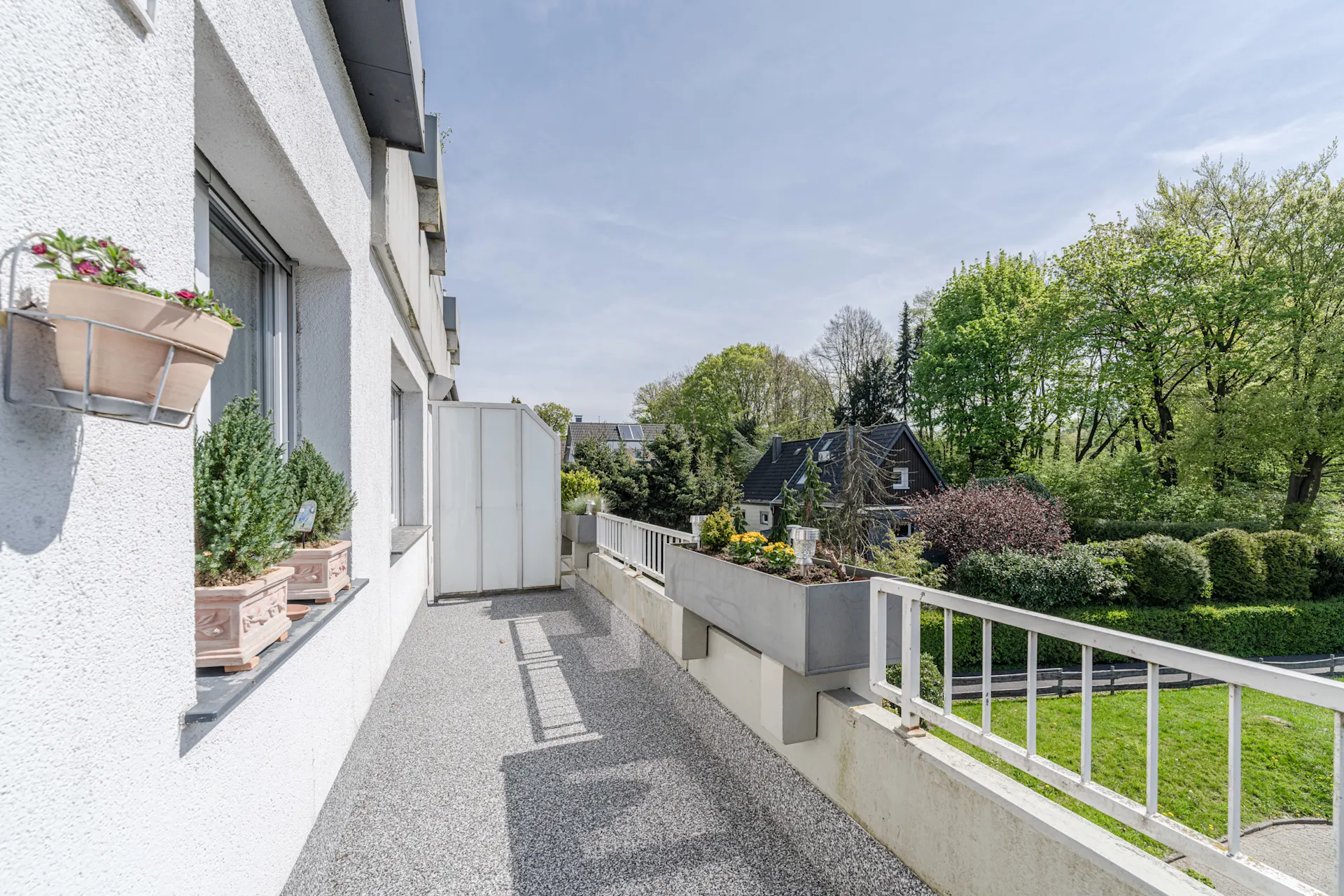 Helle Terrasse mit Blumen, Blick auf Garten und Nachbarhaus, umgeben von Bäumen.