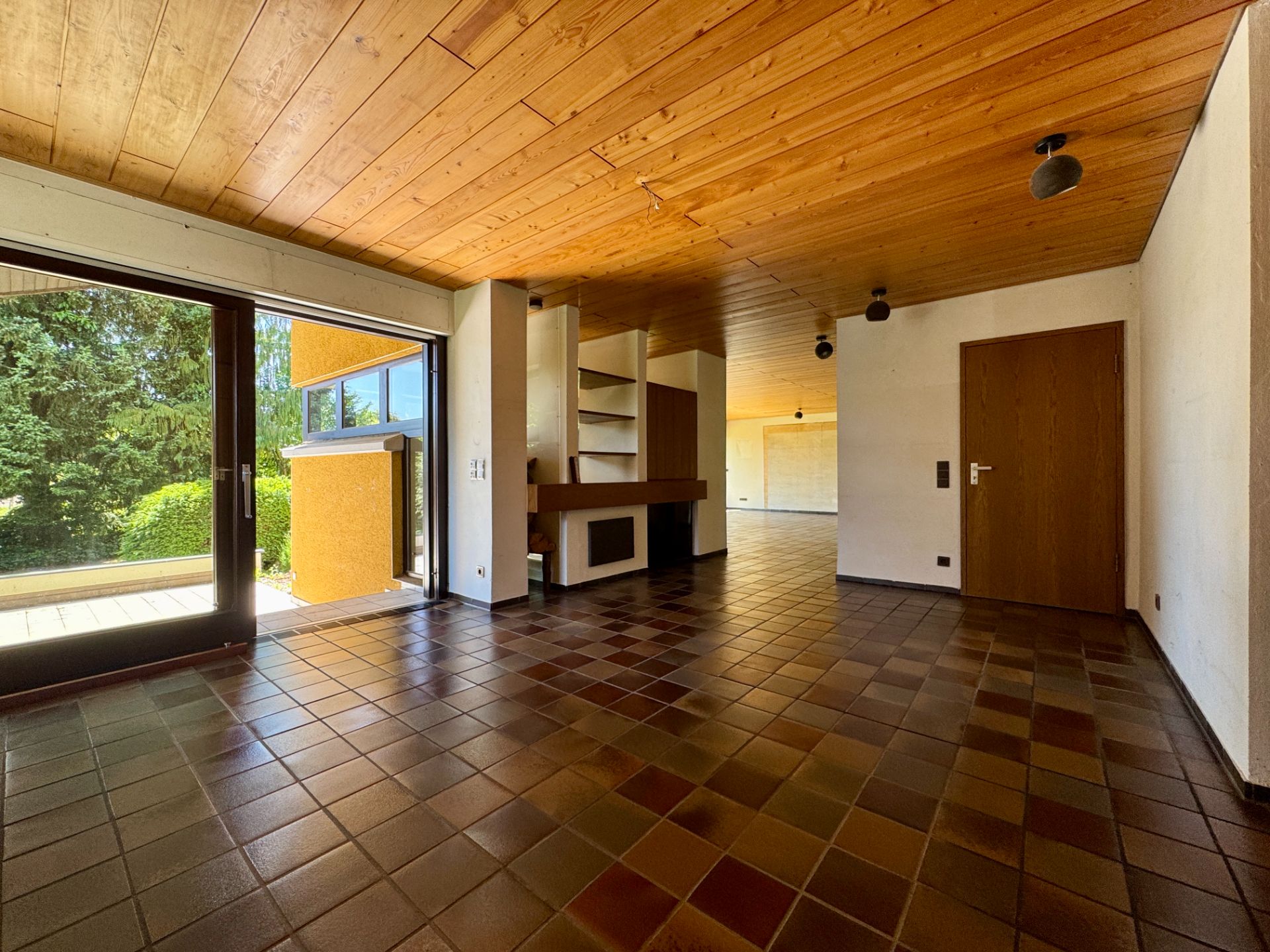Wohnzimmer mit Fliesenboden, Holzdecke, großen Fenstern und Zugang zur Terrasse.