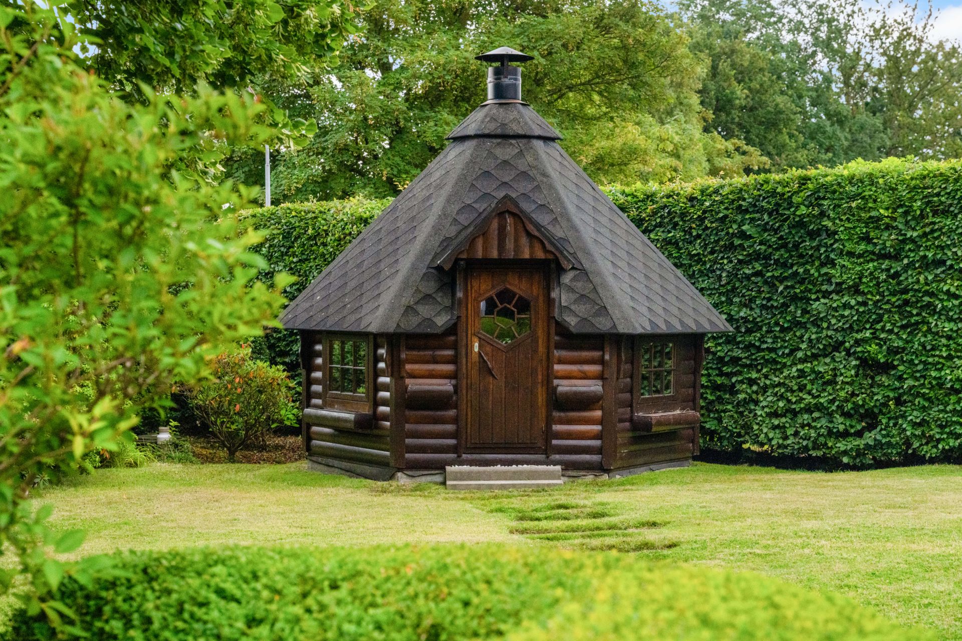 Kleines, dunkelbraunes Holzhaus mit schrägem Dach im Garten, umgeben von grüner Hecke.
