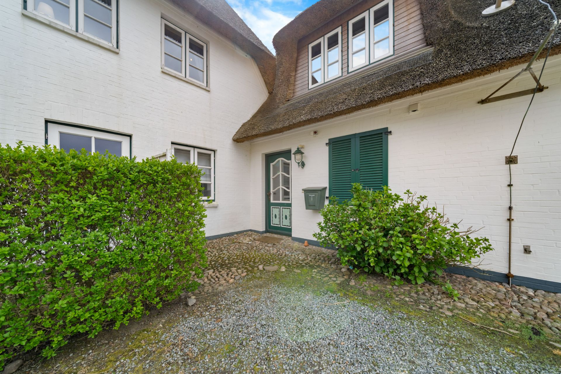 Eingangsbereich eines weißen Hauses mit grüner Tür, Fenstern und Sträuchern davor.