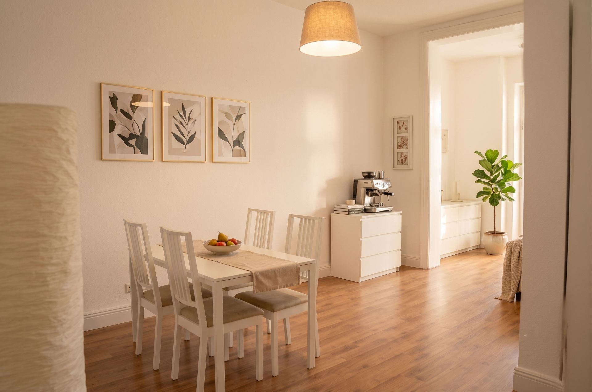 Helles Esszimmer mit Holzboden, weißem Tisch und vier Stühlen, Pflanzen und Wandbildern.