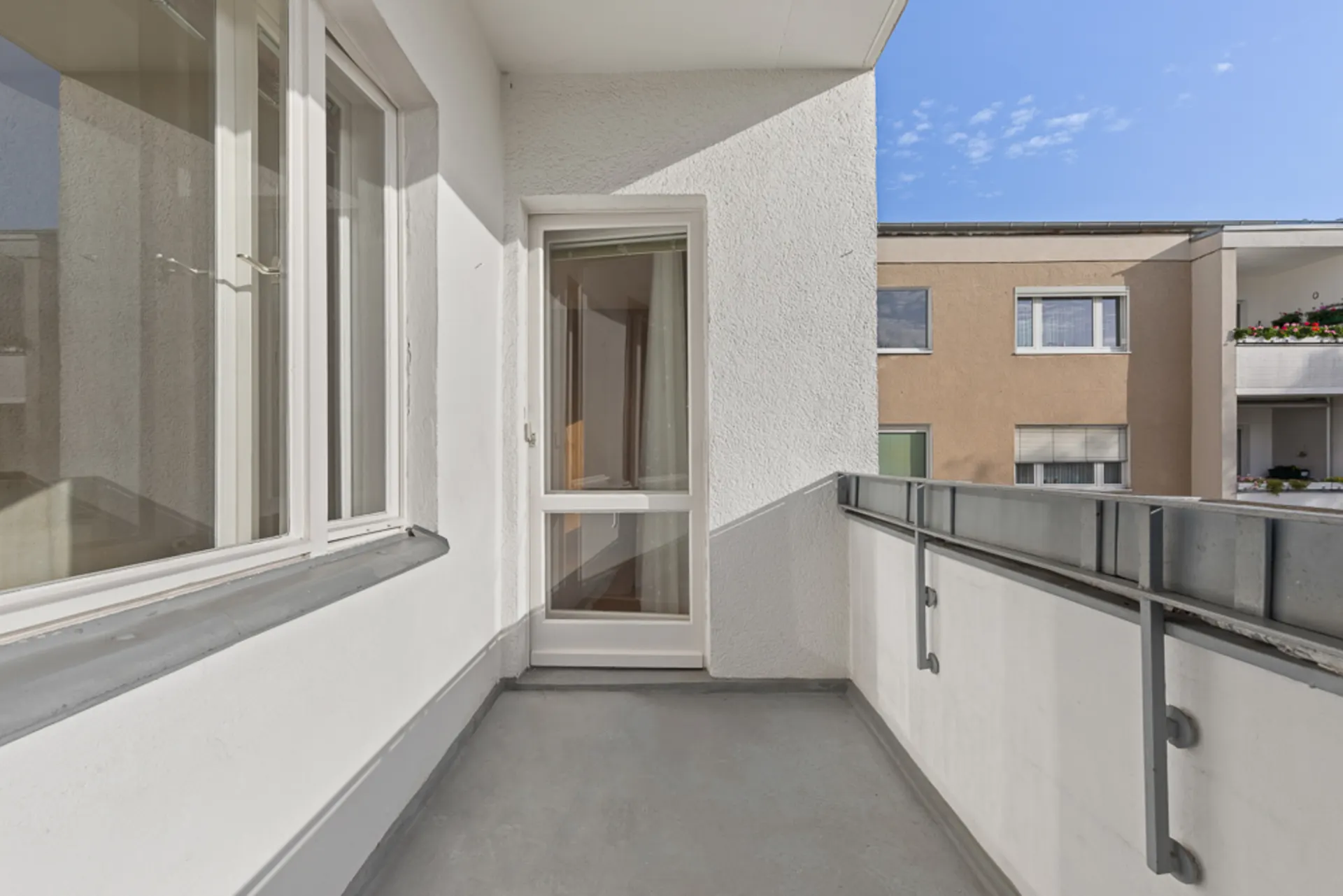 Balkon mit weißer Wand, Fenster und Glastür, Blick auf benachbarte Wohngebäude.