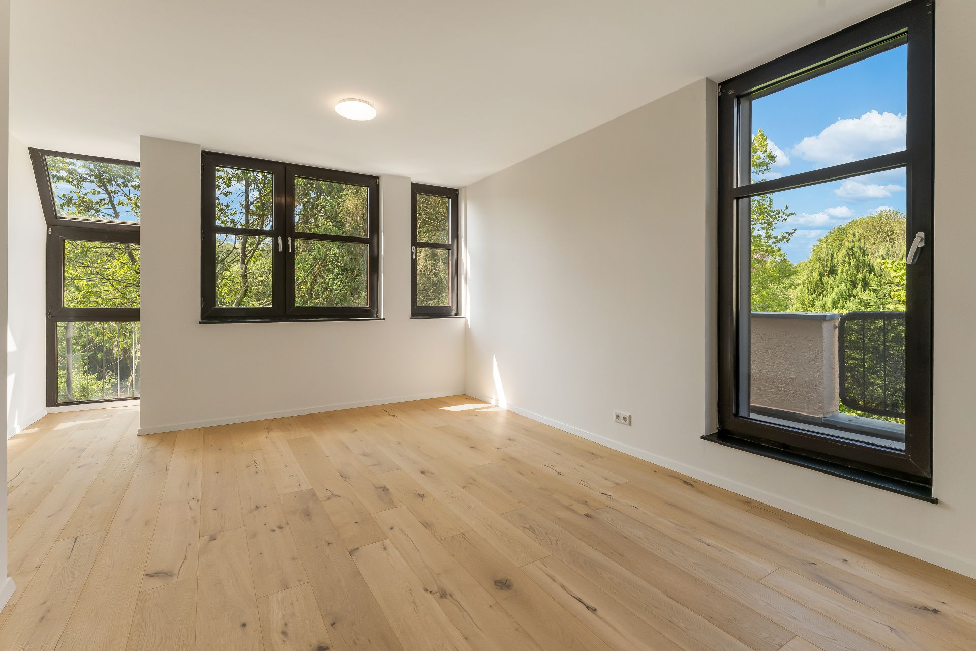 Helles Zimmer mit Holzfußboden und großen Fenstern mit Blick ins Grüne.
