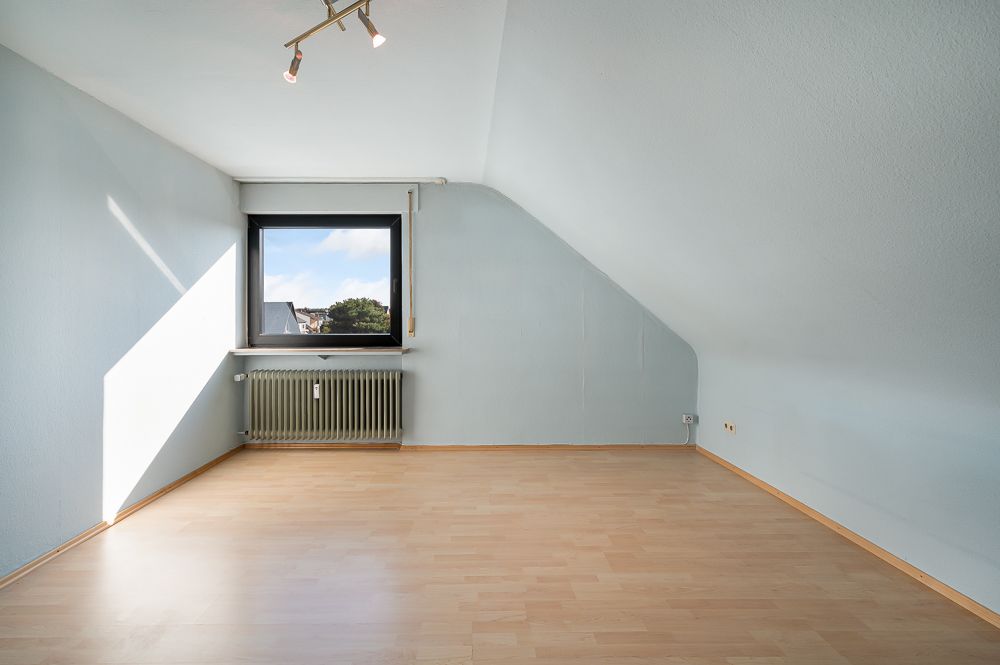 Leeres Zimmer mit Dachschräge, Fenster und Heizkörper, hellem Holzfußboden und weißer Wand.