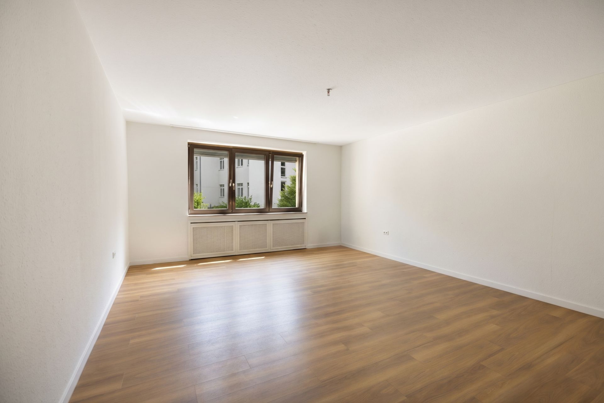 Helles, leeres Zimmer mit Holzfußboden und großem Fenster, Blick auf Nachbargebäude.
