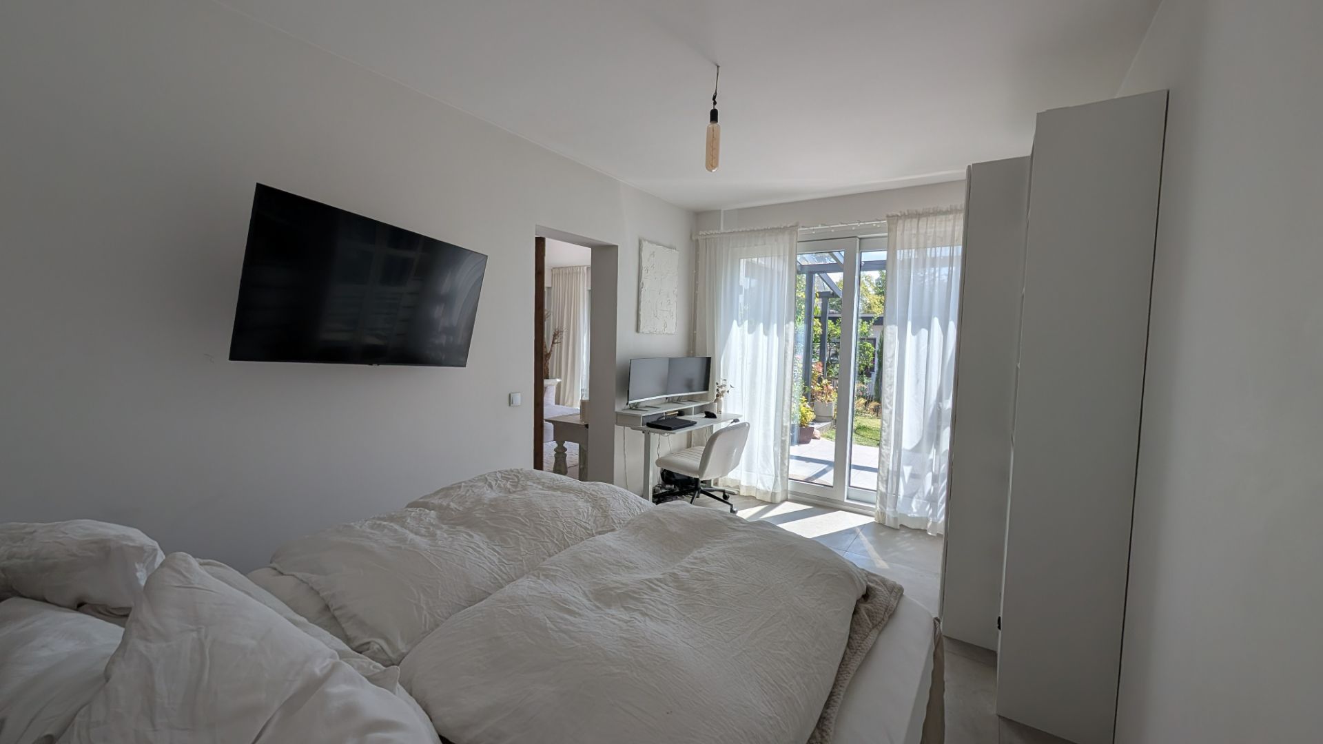 Helles Schlafzimmer mit Doppelbett, TV, Schreibtisch und Zugang zur Terrasse.