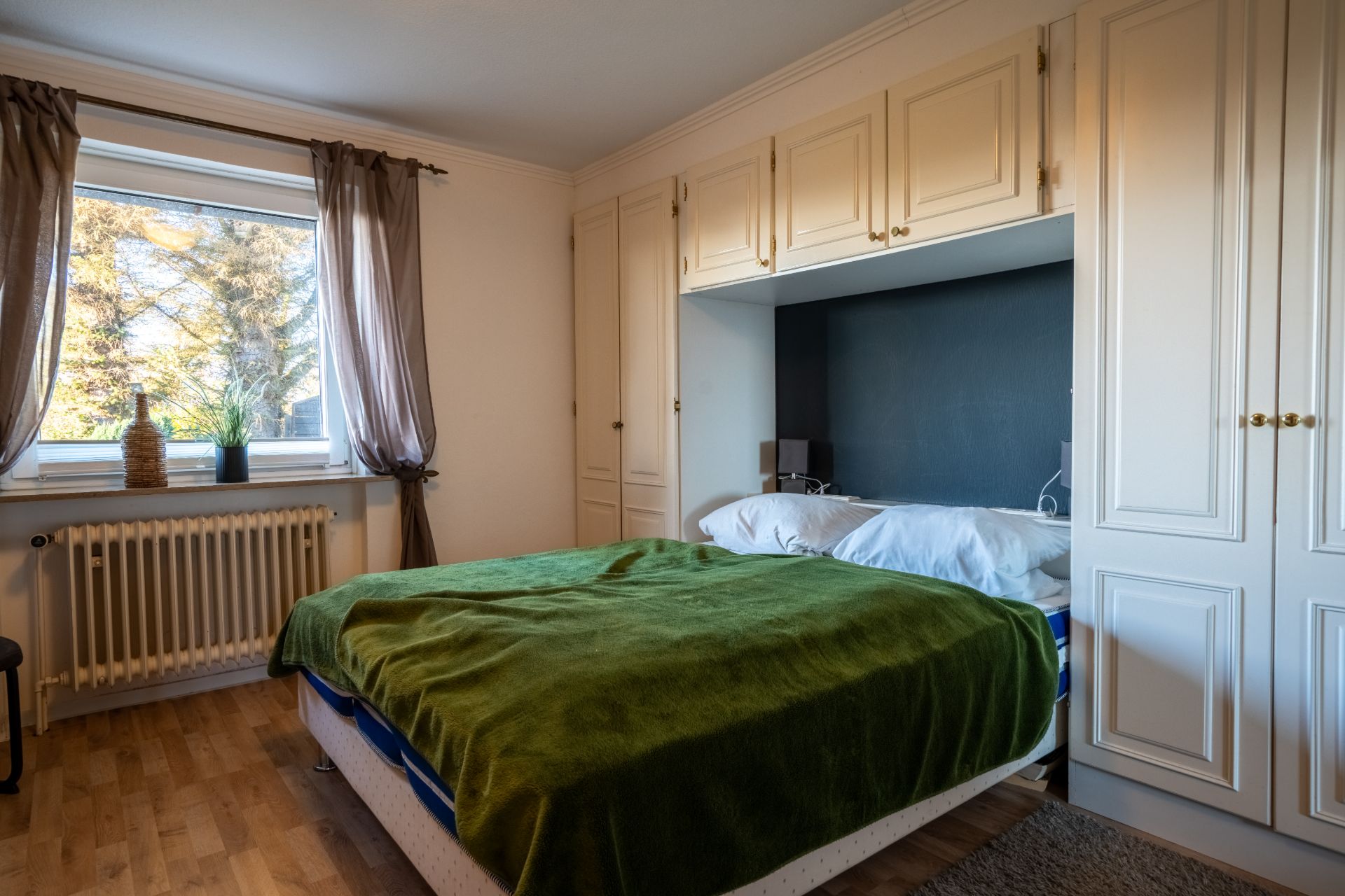 Schlafzimmer mit Bett, grüner Decke, Einbauschränken und Fenster mit Gardinen.