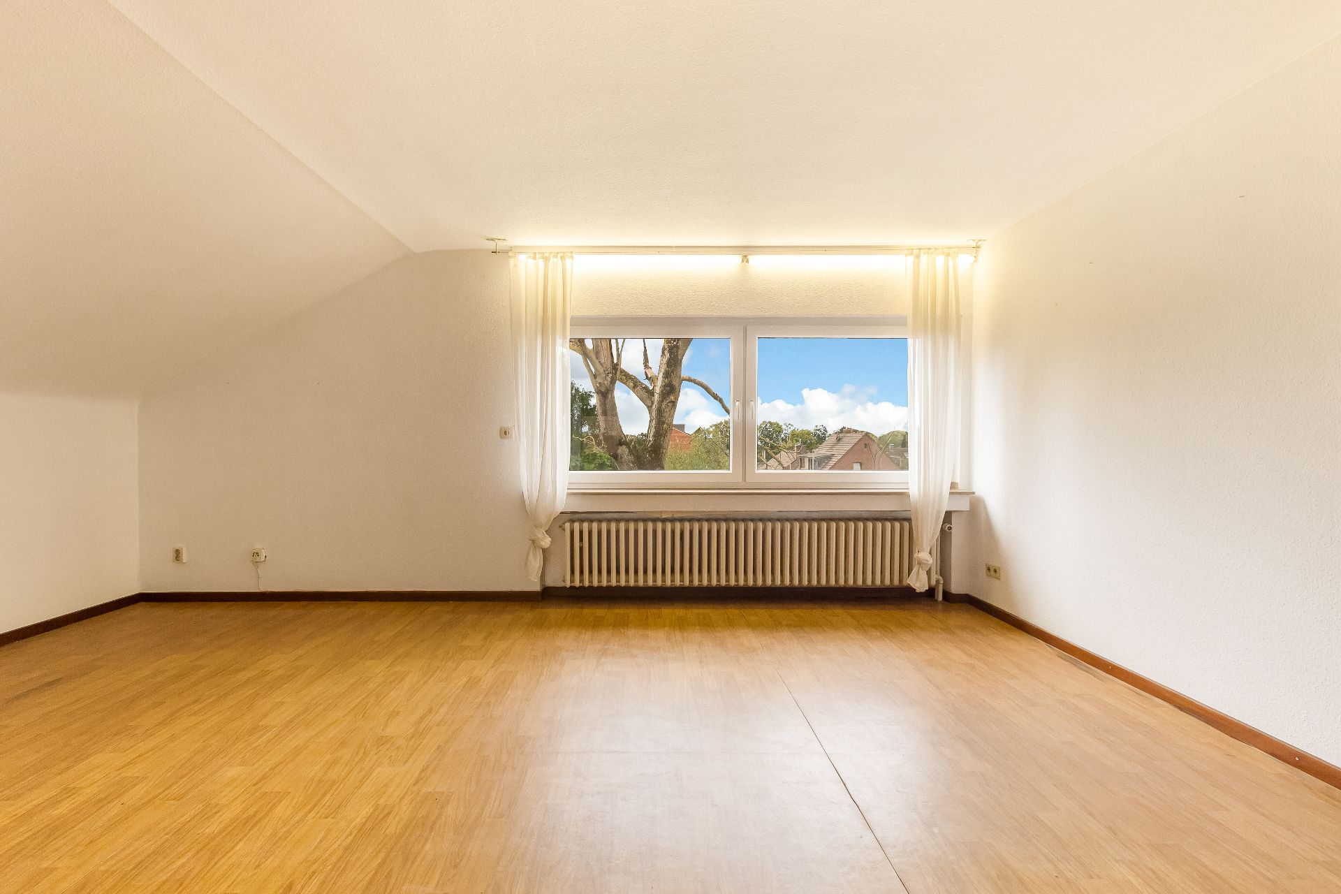 Leerer Raum mit Holzfußboden, großem Fenster, weißen Vorhängen und Heizung darunter.
