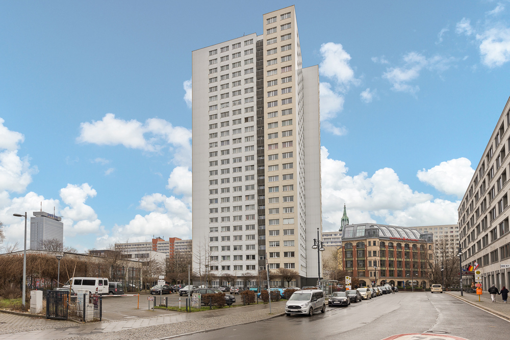 Hohes Wohnhaus mit vielen Fenstern, umgeben von Autos auf der Straße unter blauem Himmel.