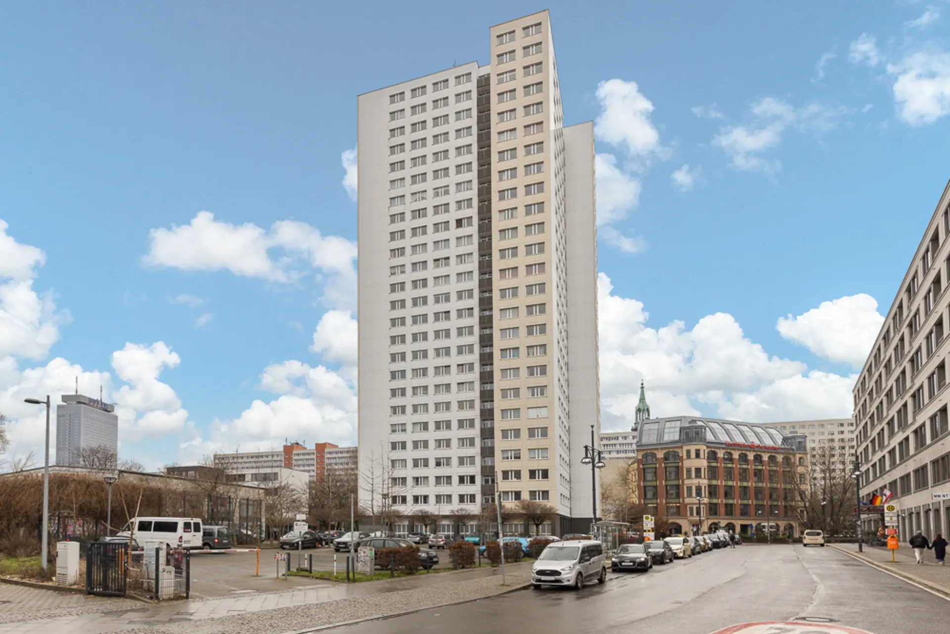 Hohes Wohnhaus mit vielen Fenstern, umgeben von Autos auf der Straße unter blauem Himmel.