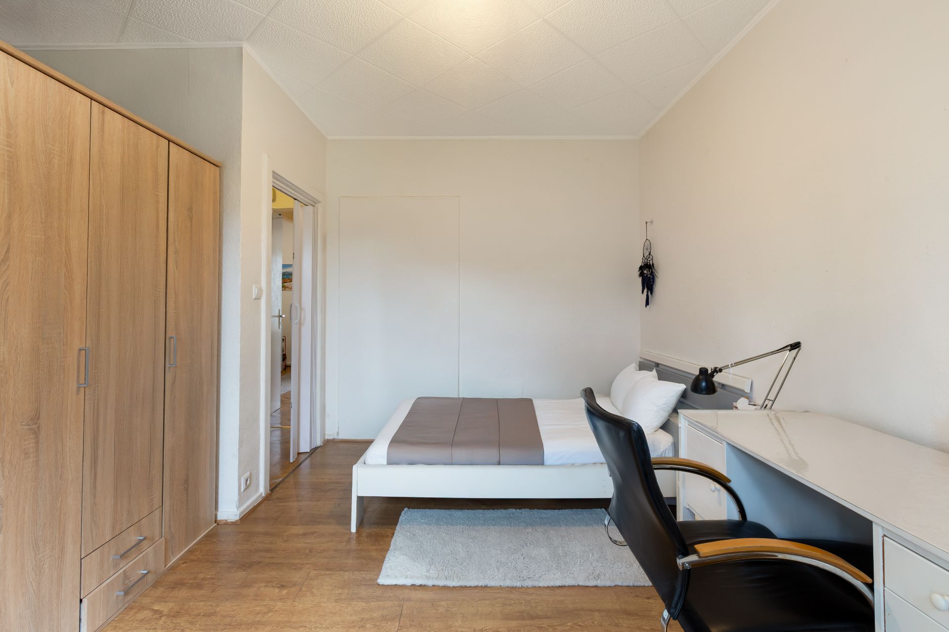 Helles Schlafzimmer mit Bett, Schreibtisch, Stuhl und Kleiderschrank auf Holzboden.