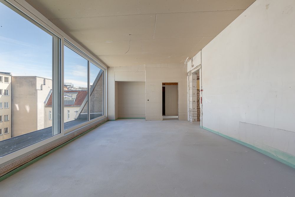 Leerer, unfertiger Raum mit großen Fenstern und Blick auf benachbarte Gebäude.