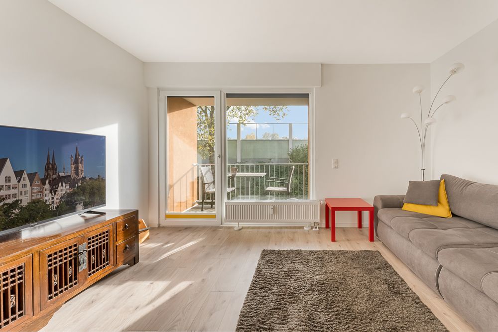 Helles Wohnzimmer mit Sofa, rotem Tisch, Fernseher und Zugang zu einem Balkon mit Stühlen.