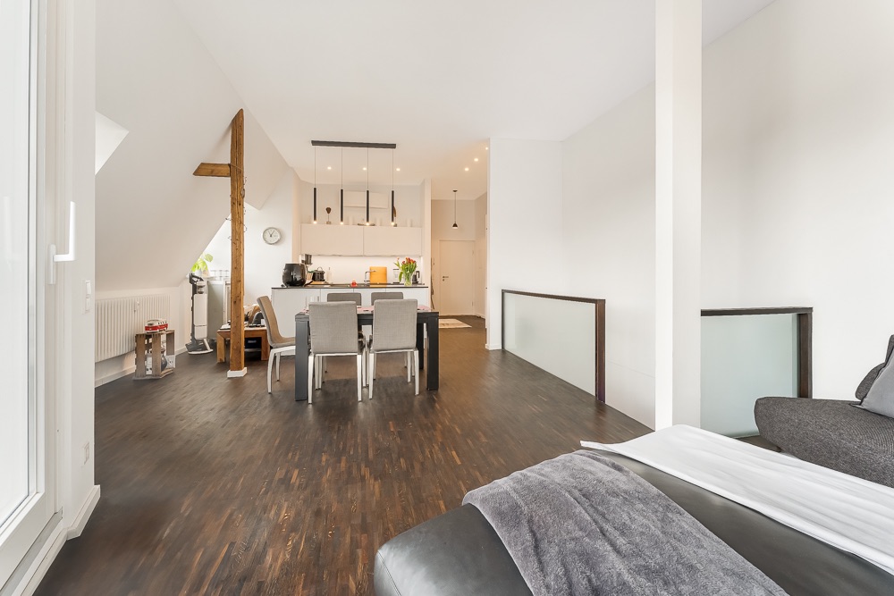 Helles Wohnzimmer mit dunklem Holzboden, Esstisch, Küche und Sofa im Vordergrund.